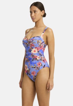 Model poseert in een bloemen badpak met een blauwe achtergrond, met een ruchesontwerp en brede schouderbanden.