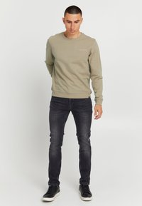 Felpa verde oliva con scollo rotondo, polsini e orlo a costine; abbinata a jeans scuri slim fit e sneakers nere. Dettaglio logo semplice sul petto.