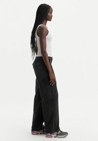 Pantaloni cargo neri con tasche laterali e una vestibilità rilassata, abbinati a una canotta bianca aderente e scarpe sportive con dettagli in rosa e grigio.