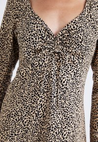 Robe à manches longues en tissu beige avec imprimé léopard noir. Présente un décolleté en V profond et un détail froncé au centre qui maintient le tissu.