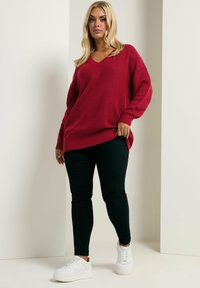 Roter gerippter Strickpullover mit V-Ausschnitt und oversized Schnitt, kombiniert mit schwarzen Slim-Fit-Jeans und weißen Sneakers. Der Look zeichnet sich durch lange Ärmel aus.