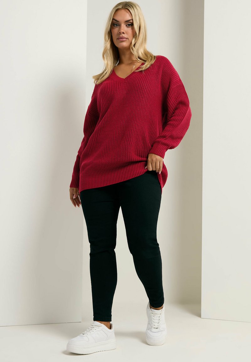 Roter gerippter Strickpullover mit V-Ausschnitt und oversized Schnitt, kombiniert mit schwarzen Slim-Fit-Jeans und weißen Sneakers. Der Look zeichnet sich durch lange Ärmel aus.