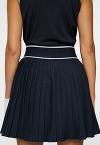 Nainen, joka pukeutuu mustaan hihattomaan paitaan ja mustaan taittokuosiseen hameeseen, jossa on leveä raidallinen vyötärönauha, kuvattu takaa.