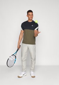 Camisa de manga corta en bloques de color azul marino y verde oliva, con detalles en blanco; combinada con pantalones gris claro y una raqueta de tenis, sosteniendo una pelota.