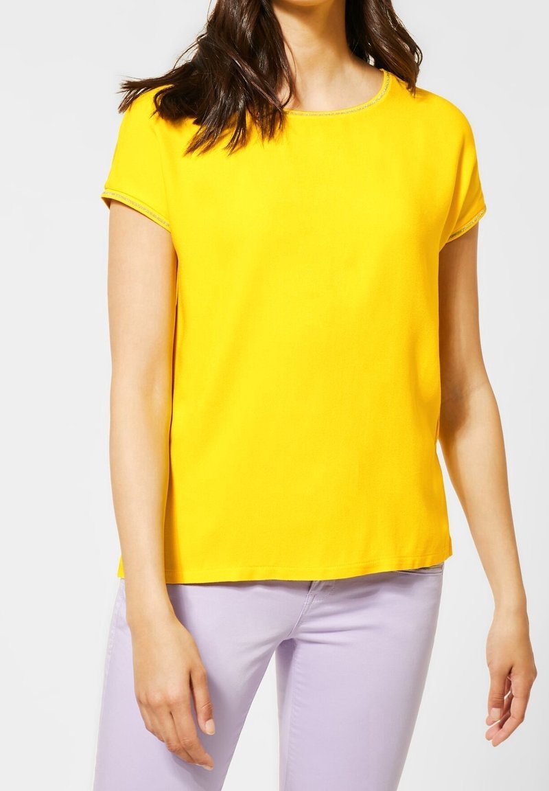 Žlutý top s krátkým rukávem a kulatým výstřihem s kontrastním lemováním. Hladká textura. Spárováno s světle fialovými kalhotami.