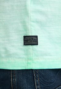 Hellgrünes Baumwollhemd mit einem schwarzen Etikett unten, das das Logo "PETROL IND & CO." zeigt. Der Stoff hat eine weiche Textur.