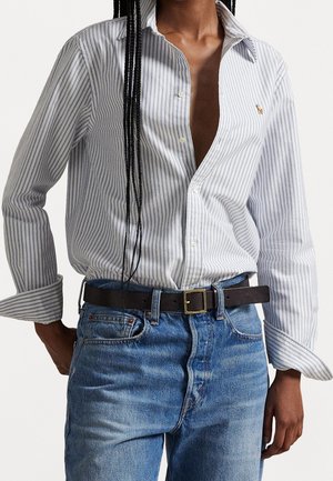 Personne portant une chemise blanche et bleue à rayures partiellement déboutonnée, rentrée dans un jean bleu avec une ceinture noire, debout devant un fond uni clair.