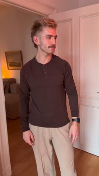 Uomo con capelli castano chiaro, baffi e orecchini, che indossa una maglietta nera a maniche lunghe e pantaloni beige, in piedi in una porta all'interno di una stanza.