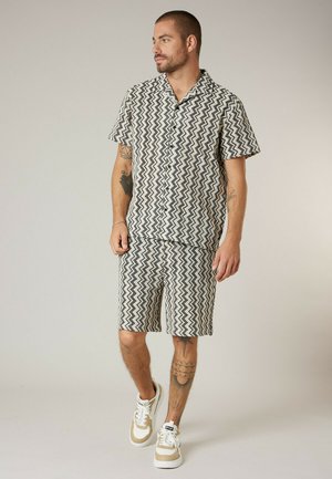 Homme aux cheveux courts et tatouages, portant une chemise à manches courtes à motif zigzag noir et blanc et un short assorti, marchant avec des baskets beige et blanc.