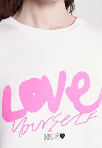 Camiseta blanca de algodón con letras en neón rosa que dicen "AMOR" y "A TI MISMO" con un acento de corazón negro y una textura suave.