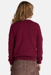 Bordeaux stickad tröja med långa ärmar, ribbade muddar och nederkant. Bakvy som visar det texturerade tyget. Bärs över en ljusrosa krage.