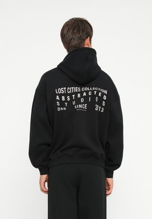 Svart hettegenser med stor trykt tekst på ryggen, med en blanding av store bokstaver. Den har en hette med snøring og ribbestrikkede mansjetter.