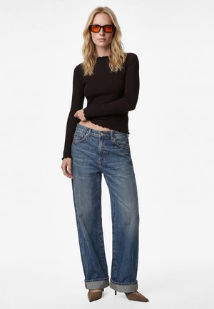 Kvinde iført orangefarvede solbriller, sort langærmet top, blå jeans med brede ben og opbuk, samt brune spidse højhælede sko, stående.