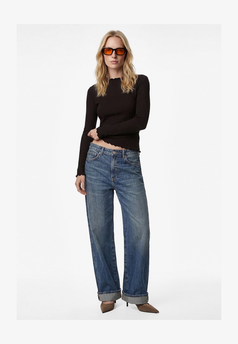 Femme portant des lunettes de soleil à teinte orange, un haut noir à manches longues, un jean bleu à jambes larges retroussé, et des chaussures marron à talons aiguilles pointues, debout.