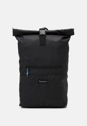 Sac à dos noir à fermeture roulante, poche avant zippée, tirette bleue et fermeture à boucle sur un fond uni clair.