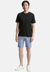 Zwarte katoenen t-shirt, lichtblauwe opgerolde shorts en witte sneakers. Model staat tegen een effen achtergrond en toont casual zomeroutfit.
