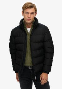 Chaqueta acolchada negra con un cuello alto, diseño acolchado y puños de canalé. Lleva puesta encima de un suéter verde de punto trenzado, combinado con jeans oscuros.
