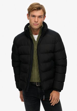 Chaqueta acolchada negra con un cuello alto, diseño acolchado y puños de canalé. Lleva puesta encima de un suéter verde de punto trenzado, combinado con jeans oscuros.