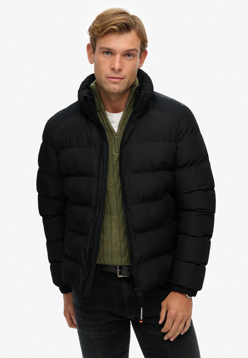 Chaqueta acolchada negra con un cuello alto, diseño acolchado y puños de canalé. Lleva puesta encima de un suéter verde de punto trenzado, combinado con jeans oscuros.
