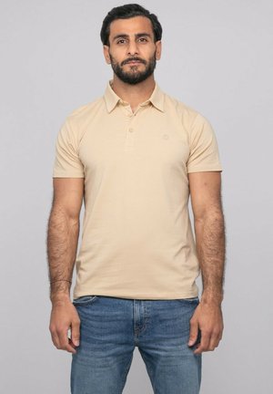Homme aux cheveux foncés et à la barbe, portant un polo beige et un jean bleu, debout devant un fond uni gris clair.