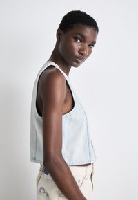 Lyseblå denim crop top med brede skulderstropper og rå kant. Tøyet har en jevn tekstur, sammen med kremfargede bukser med blå detaljer.