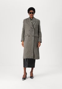 Abrigo de sarga de doble botonadura en tela gris y negra, con amplios solapas y dos bolsillos frontales, combinado con una falda de cuero y tacones.