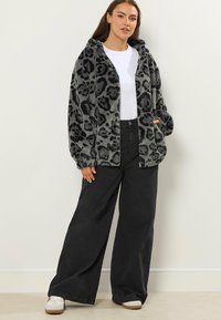 Haut à capuche en fausse fourrure grise avec des taches noires de léopard, doté d'un zip et de deux poches latérales, associé à un jean noir à jambes larges et à des baskets blanches.