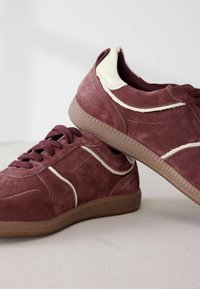 Paire de baskets en suède bordeaux avec des accents beiges et des semelles en caoutchouc marron, posées sur une surface claire.