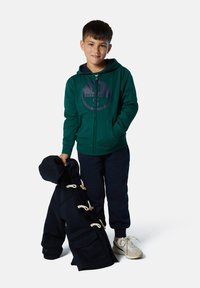 Felpa verde con zip e logo circolare, abbinata a pantaloni jogger blu navy. Un cappotto blu navy con dettagli in corda è appoggiato sul braccio. Indossa scarpe da ginnastica bianche.
