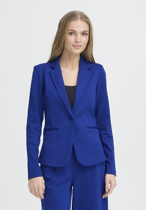 Mujer joven con cabello largo que lleva una blazer azul real ajustada y pantalones a juego, de pie contra un fondo claro y liso.