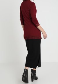Bordowa bluzka z długim rękawem z koronkową lamówką u dołu, zestawiona z czarną spódnicą i botkami, podkreślająca dopasowaną sylwetkę.