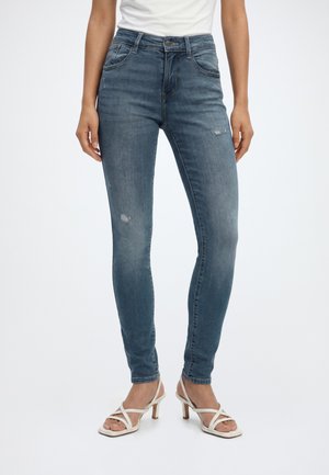 Femme portant un jean skinny bleu légèrement usé et des sandales blanches à talons, debout devant un fond uni.