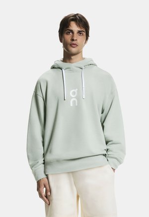 Giovane uomo con felpa con cappuccio verde chiaro con logo bianco e coulisse, abbinata a pantaloncini color crema, in piedi davanti a uno sfondo bianco.