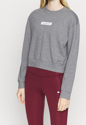 Osoba ubrana w szarą bluzę Calvin Klein i bordowe legginsy z małym logo, stojąca na tle jednolitego jasnego tła.