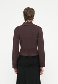 Pessoa com cabelo castanho curto, vestindo um suéter justo em tons de castanho escuro e uma saia preta, em pé contra um fundo branco liso.