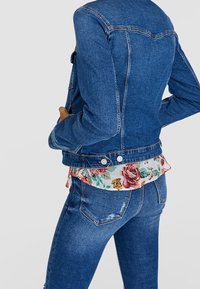 Chaqueta de mezclilla en azul oscuro con un diseño ajustado, que presenta dos botones a presión en el dobladillo y una tela floral asomando por debajo de la chaqueta.