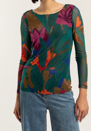 Vrouw draagt een lange mouw, aansluitend mesh top met een kleurrijk abstract bloemmotief in groen, paars, oranje en bruin, gecombineerd met een blauwe spijkerbroek.