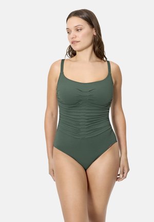 Mujer con un bañador de una pieza verde con detalles fruncidos, de pie frente a un fondo gris claro liso, mirando hacia un lado.