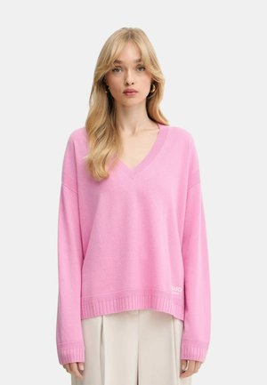 Donna con lunghi capelli biondi che indossa un ampio maglione rosa a V e pantaloni beige chiari, in piedi contro uno sfondo chiaro e uniforme.