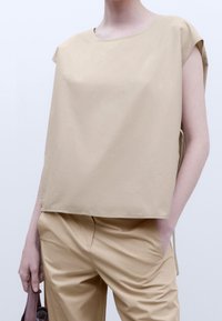 Top beige oversize con maniche corte, texture liscia, scollatura rotonda e lacci laterali. Abbinato a pantaloni beige a taglio slim in tinta unita.