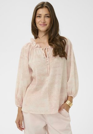 Femme aux longs cheveux bruns portant une blouse rose clair à encolure à volants et manches amples, associée à un pantalon rose clair et un bracelet manchette doré.
