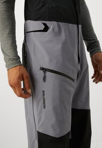 Pantaloni de schi impermeabili gri și negri, cu buzunare cu fermoar și finisaj texturat, având talie cu velcro și branding "Salomon".