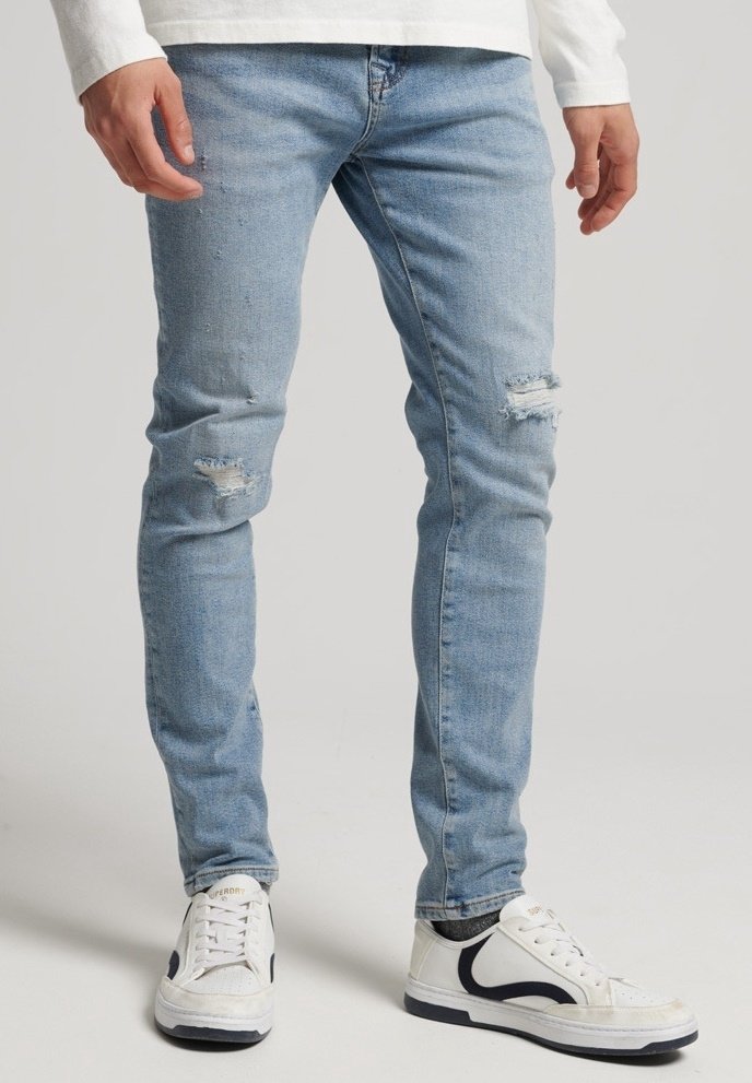 Vaqueros de mezclilla azul claro con áreas desgastadas, diseño ajustado, cintura a media altura, combinados con zapatillas blancas con acentos en marino.
