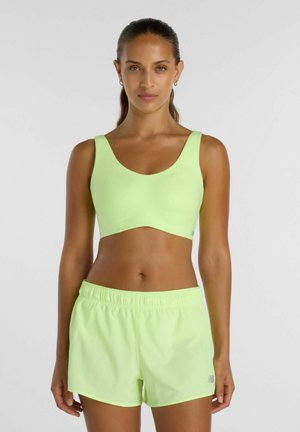 Vrouw met lang haar naar achteren gebonden, gekleed in een lichtgroene sportbeha en bijpassende shorts met elastische taille, staand tegen een effen achtergrond.