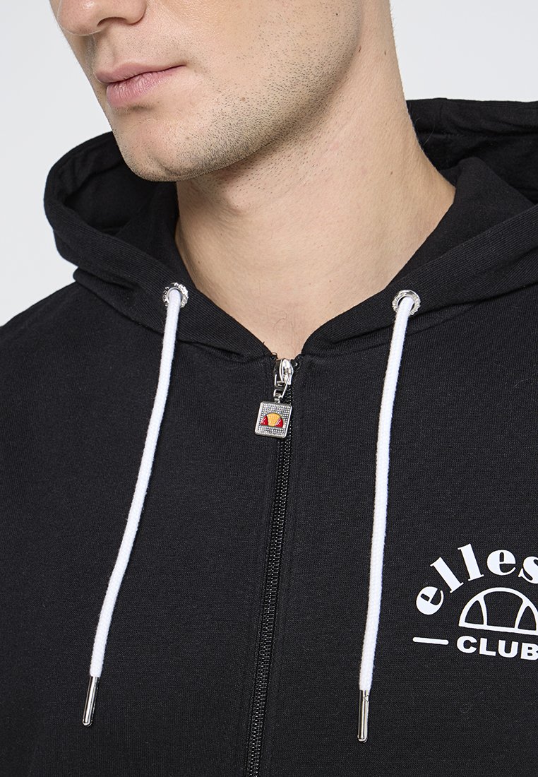 Primer plano de una persona que lleva una sudadera con cremallera negra y cordones blancos, con un logo blanco de "ellesse club" en el pecho.