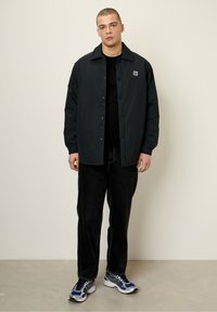 Black, lightweight jacket with a button-up front, matching collar, and logo patch. Worn over a black shirt with dark jeans and blue sneakers.