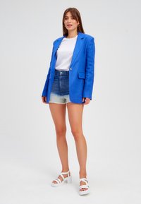 Blazer di lino blu sopra una maglietta bianca, abbinato a pantaloni di jeans corti. Sandali con tacco alto bianchi completano il look.