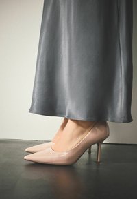 Alasti läikiva lakknahaga kõrge kontsaga kingad terava varbaosaga, kombineeritud voolava halli satiinse seelikuga. Sile tekstuur ja elegantne viimistlus.