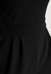 Détail d'une robe en tissu noir avec une large ceinture et des plis doux sous la taille, montrant les lignes de couture et une texture lisse.