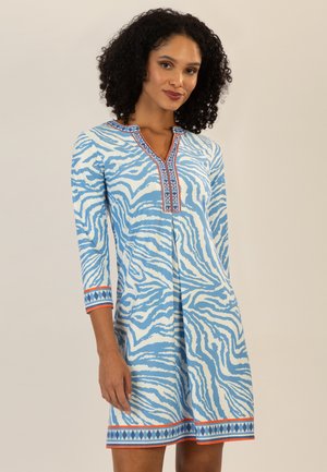 Frau mit lockigem Haar, die ein blau-weiß gestreiftes Kleid mit Zebramuster trägt, mit dreiviertellangen Ärmeln und geometrischem Besatz an Kragen, Manschetten und Saum.